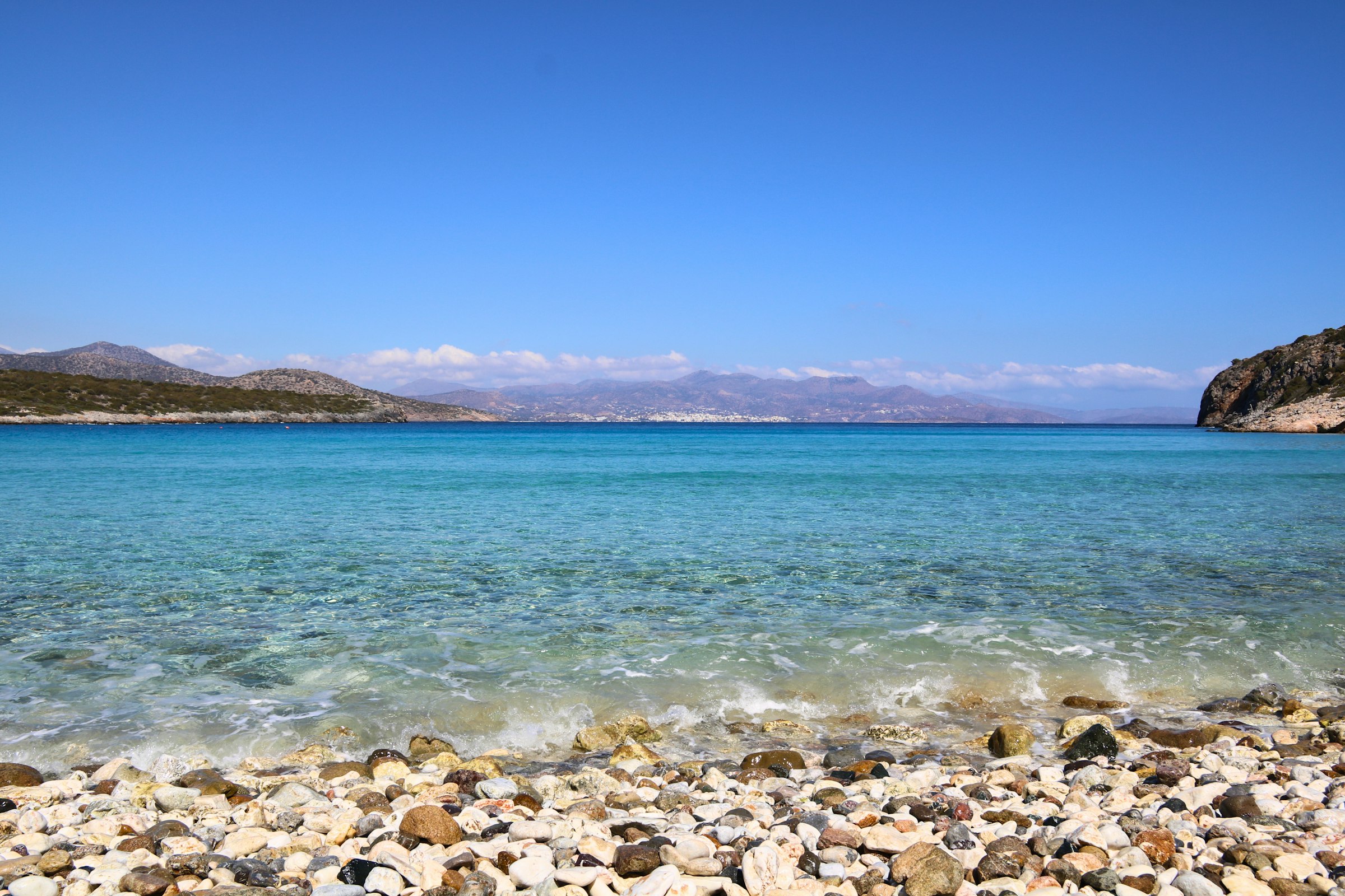 Une crique de galets clairs aux eaux turquoise cristallines, des collines arides et des montagnes au loin sous un ciel bleu — côte de la Crète.