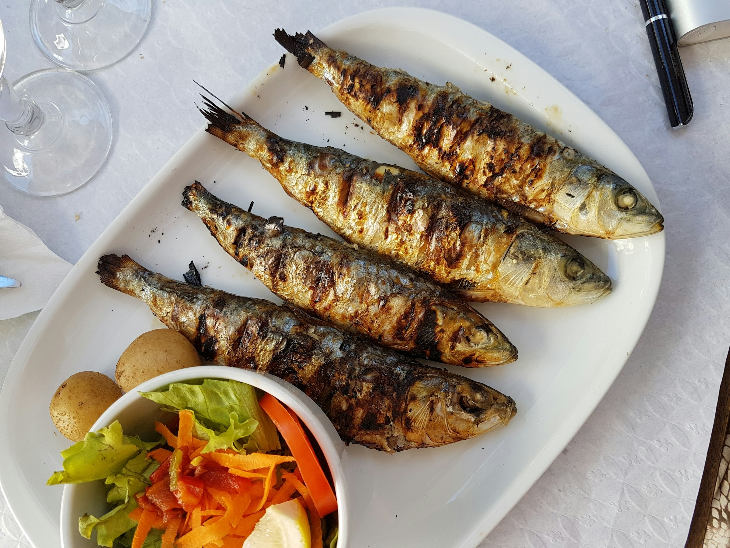 Quatre poissons entiers grillés (sardines) aux marques de grill, sur une assiette blanche, avec une petite salade et un quartier de citron.