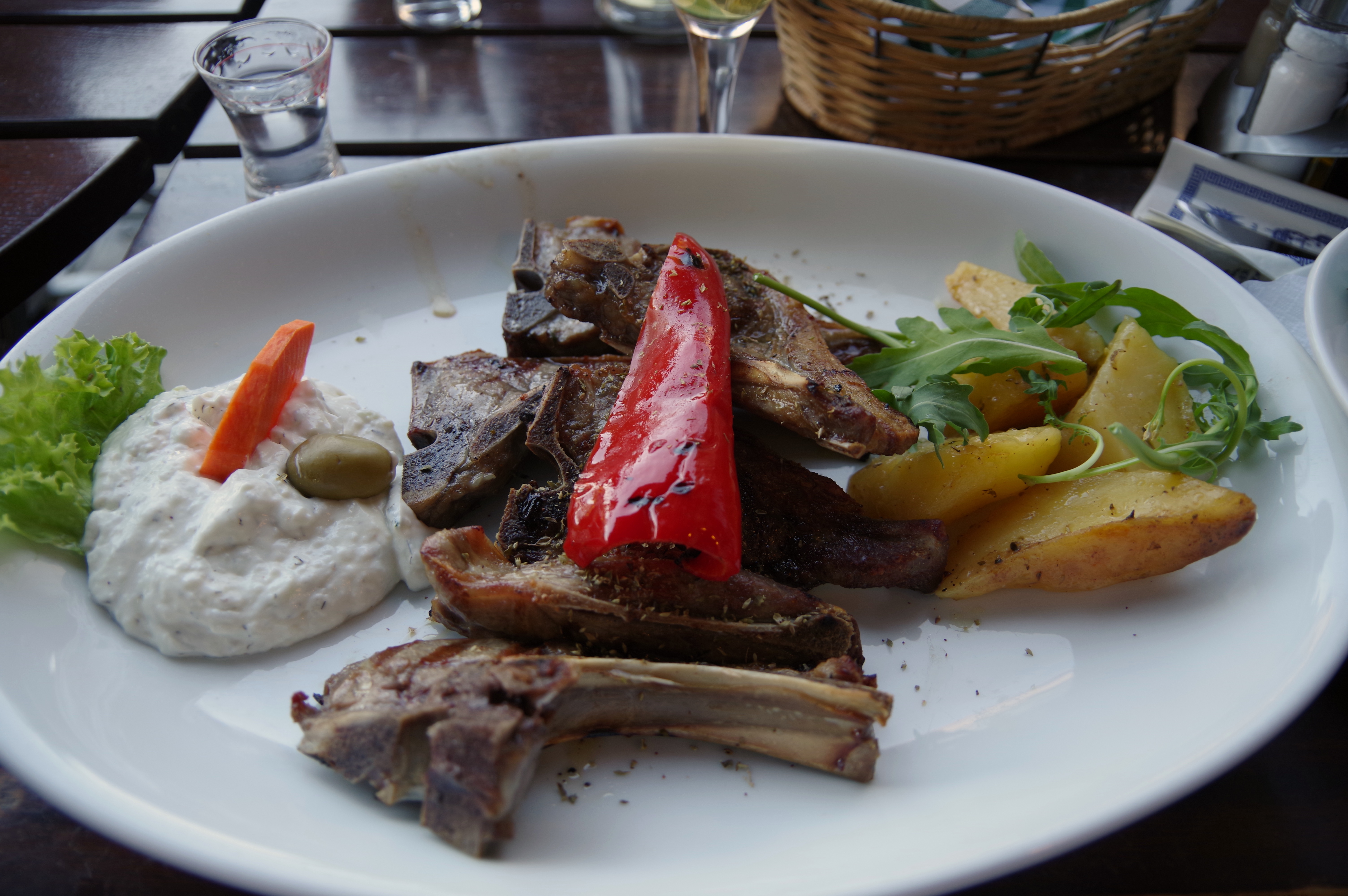 Une assiette de côtelettes d'agneau grillées (paidakia) dans une taverne grecque, avec un poivron rouge grillé, des pommes de terre rôties, de la roquette et une quenelle de trempette blanche, sur une table sombre