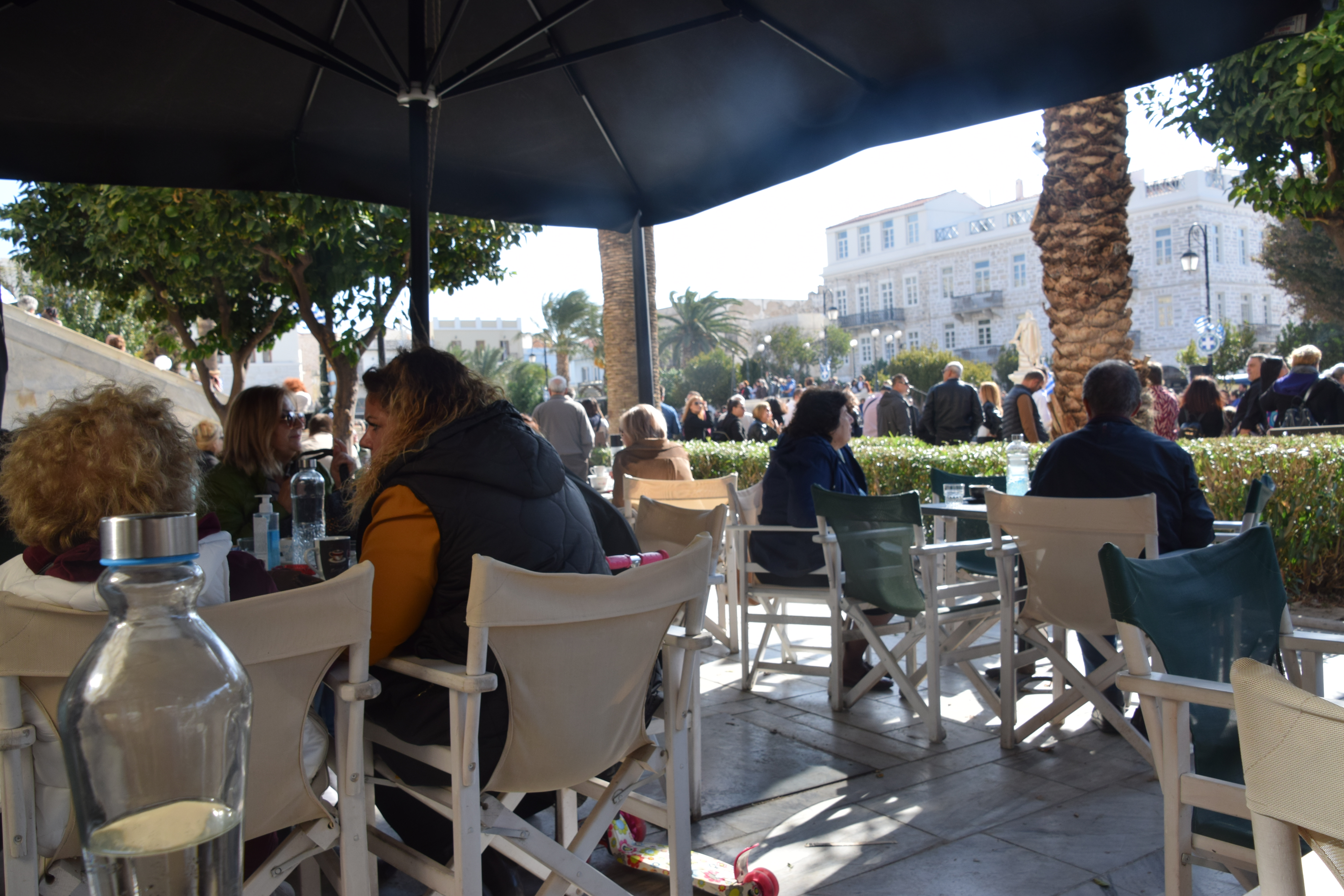 La terrasse d'un café sur la place Miaouli d'Ermoupoli : des gens attablés sous un parasol, une carafe d'eau en avant-plan, l'hôtel de ville néoclassique et la foule sur la place de marbre derrière, en lumière chaude de fin de journée