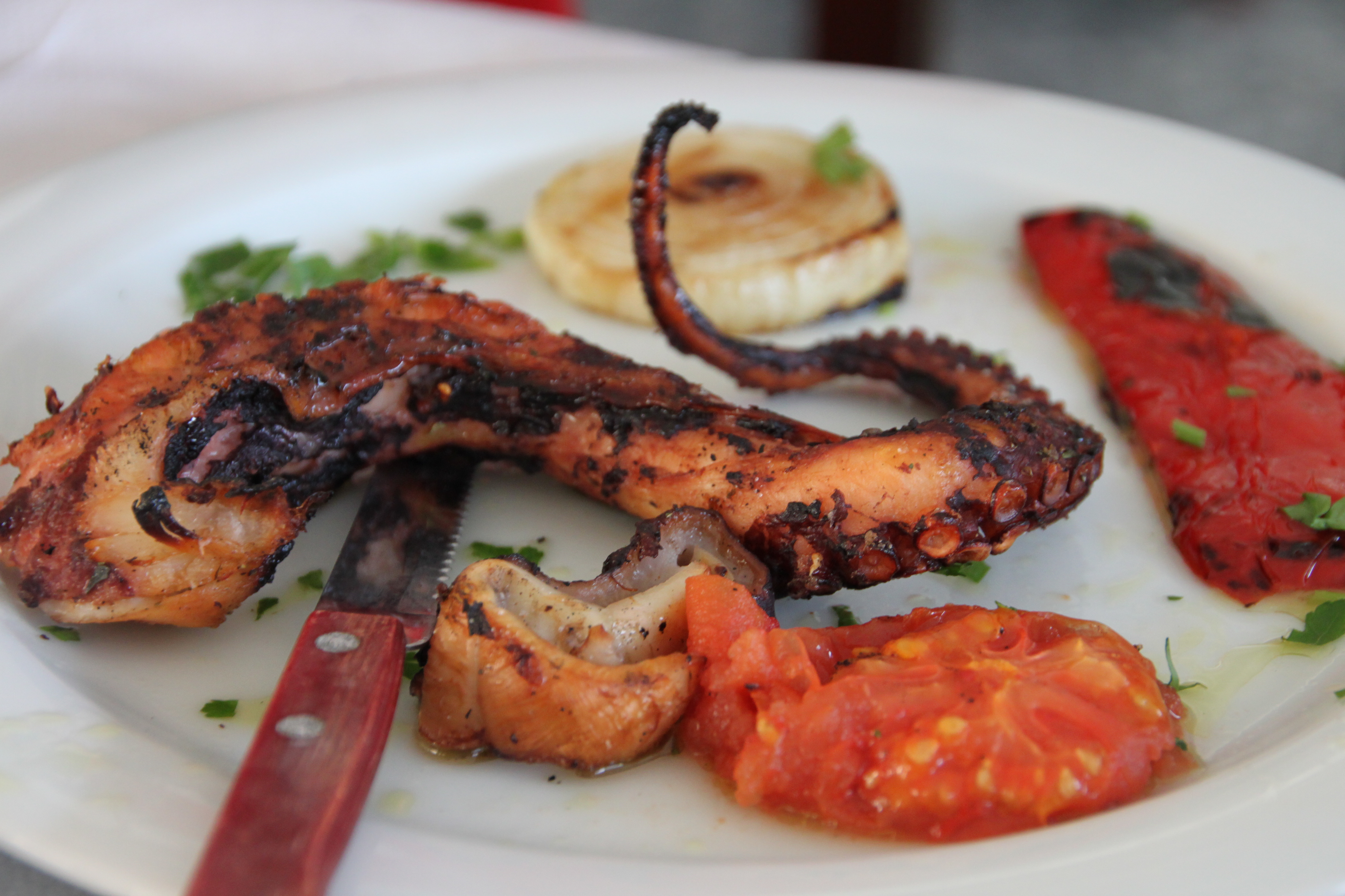 Une tentacule de poulpe grillée au charbon (chtapodi sta karvouna), dorée et charbonnée, dans une assiette avec une tranche d'oignon grillée, un poivron rouge grillé, une demi-tomate et du persil, arrosés d'huile d'olive